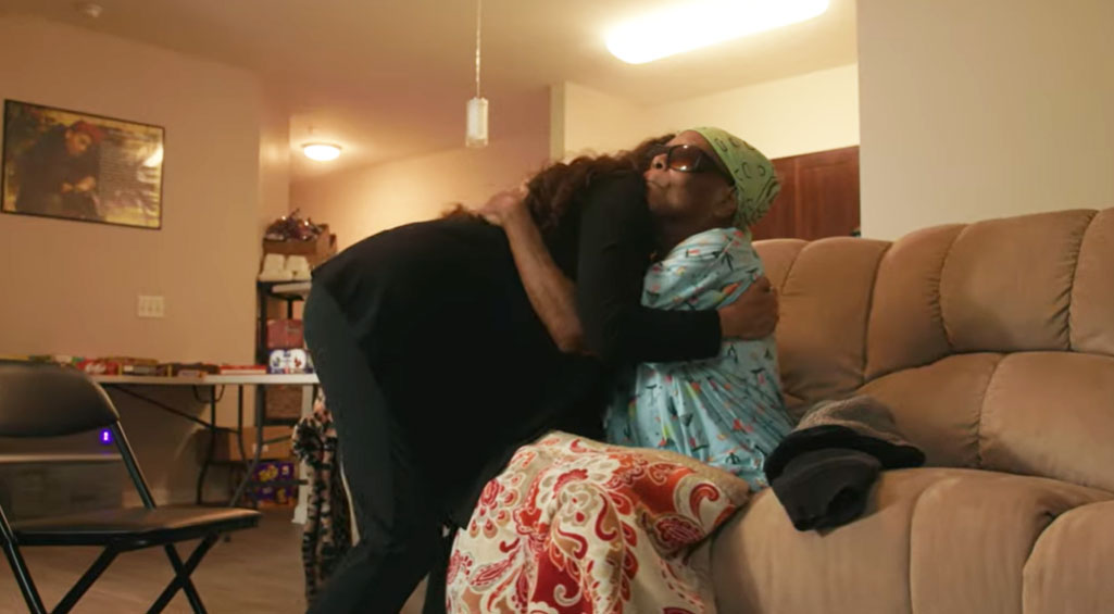Doctor leaning down hugging patient while patient is sitting on couch in their living room.
