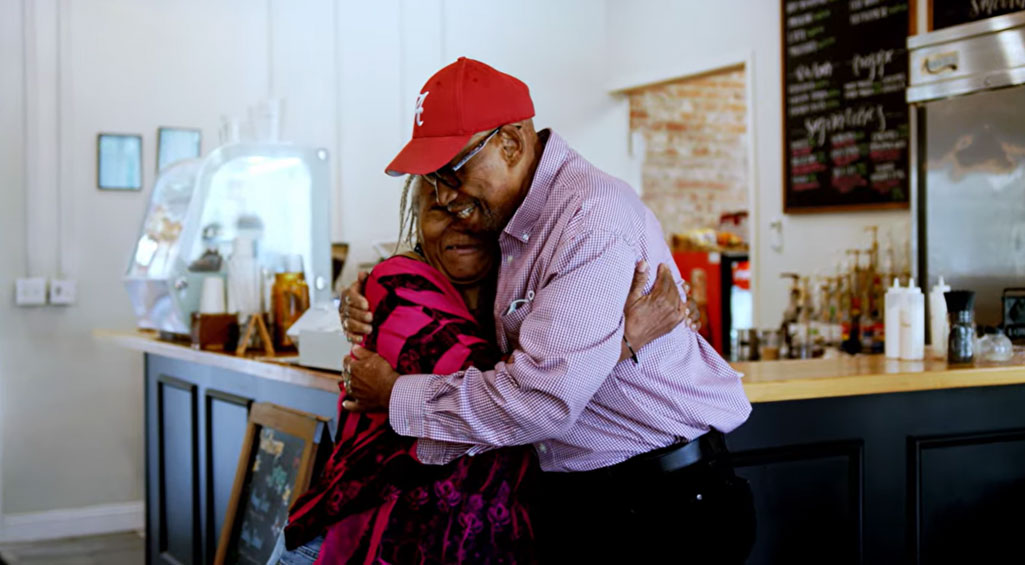 Husband and wife, standing up and hugging each other in a cafe.